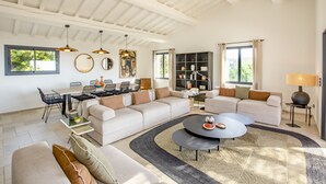 A spacious living area with a large sofa, a dining table with chairs, and a coffee table. There are pendant lights, a bookshelf, and a lamp.