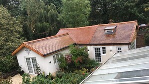 Une maison avec un toit recouvert de tuiles rouges, des murs blancs et un jardin où poussent différentes plantes, ainsi qu’une serre.