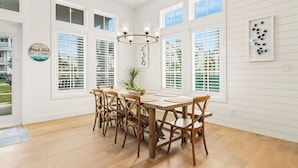 A dining area with a wooden table and chairs, large windows with blinds, and a chandelier. - Inviting Beach Home w/ Cinnamon Shore South Style & Amenities (Port Aransas)