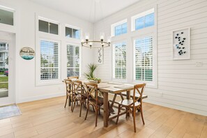 Un comedor con mesa y sillas de madera, ventanas amplias con persianas y una araña.
