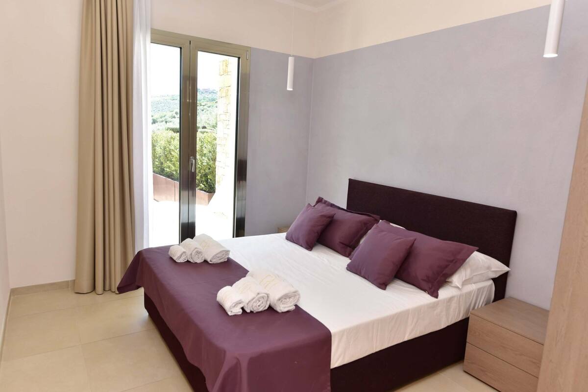 A bedroom with a large bed, a bedside table, a window with curtains, and a view of greenery outside.