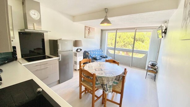 A compact kitchen with a stove, refrigerator, and a clock on the wall. There is a dining area with a table and chairs, and a window providing a view of greenery outside.