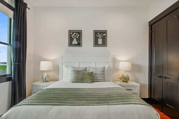 Un dormitorio ordenado con una cama grande, dos mesitas de noche, una ventana con cortinas y una puerta.
