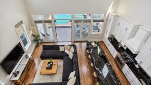 A modern living room with a black sofa, wooden coffee table, and a TV. There is a large window overlooking a pool and outdoor seating.