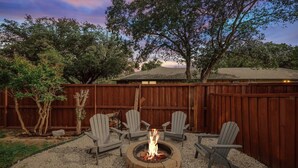 A backyard with a fire pit surrounded by Adirondack chairs, a wooden fence, and trees.