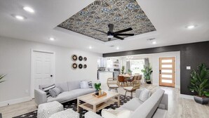 A modern living room with a sofa, coffee table, and chairs, a kitchen in the background, and a ceiling fan.
