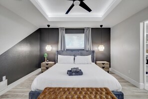 A modern bedroom with a large bed, two bedside tables, a ceiling fan, and a window with curtains.