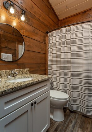 A bathroom with wooden walls, a granite countertop sink, a white toilet, and a striped shower curtain. - Walk2DowntownPF/GasFirepit/Waterfront (Pigeon Forge)