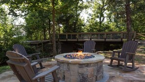 A stone fire pit surrounded by Adirondack chairs in a wooded outdoor setting. - Walk2DowntownPF/GasFirepit/Waterfront (Pigeon Forge)