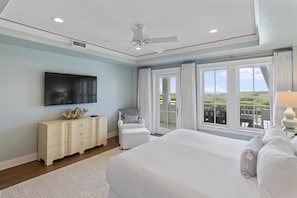 Quarto com uma cama grande, ventilador de teto, televisão, penteadeira e vista para o exterior por grandes janelas.