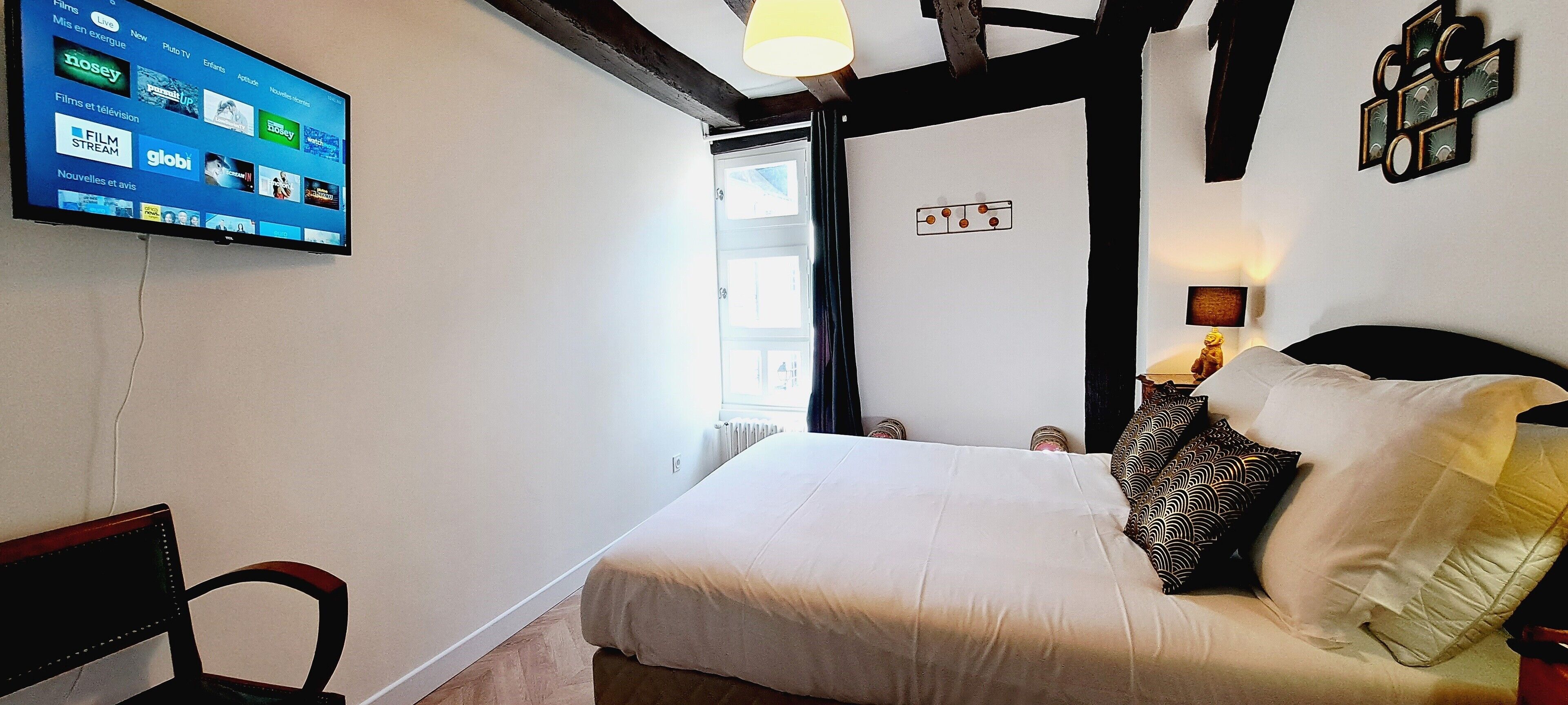 A hotel room with a bed, a chair, a TV displaying a film and TV channel interface, and a window with a view of the outside.