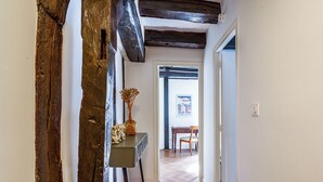 Un couloir avec des poutres apparentes, une table surmontée d’un vase, et un coin repas où l’on aperçoit une chaise et une table au fond.