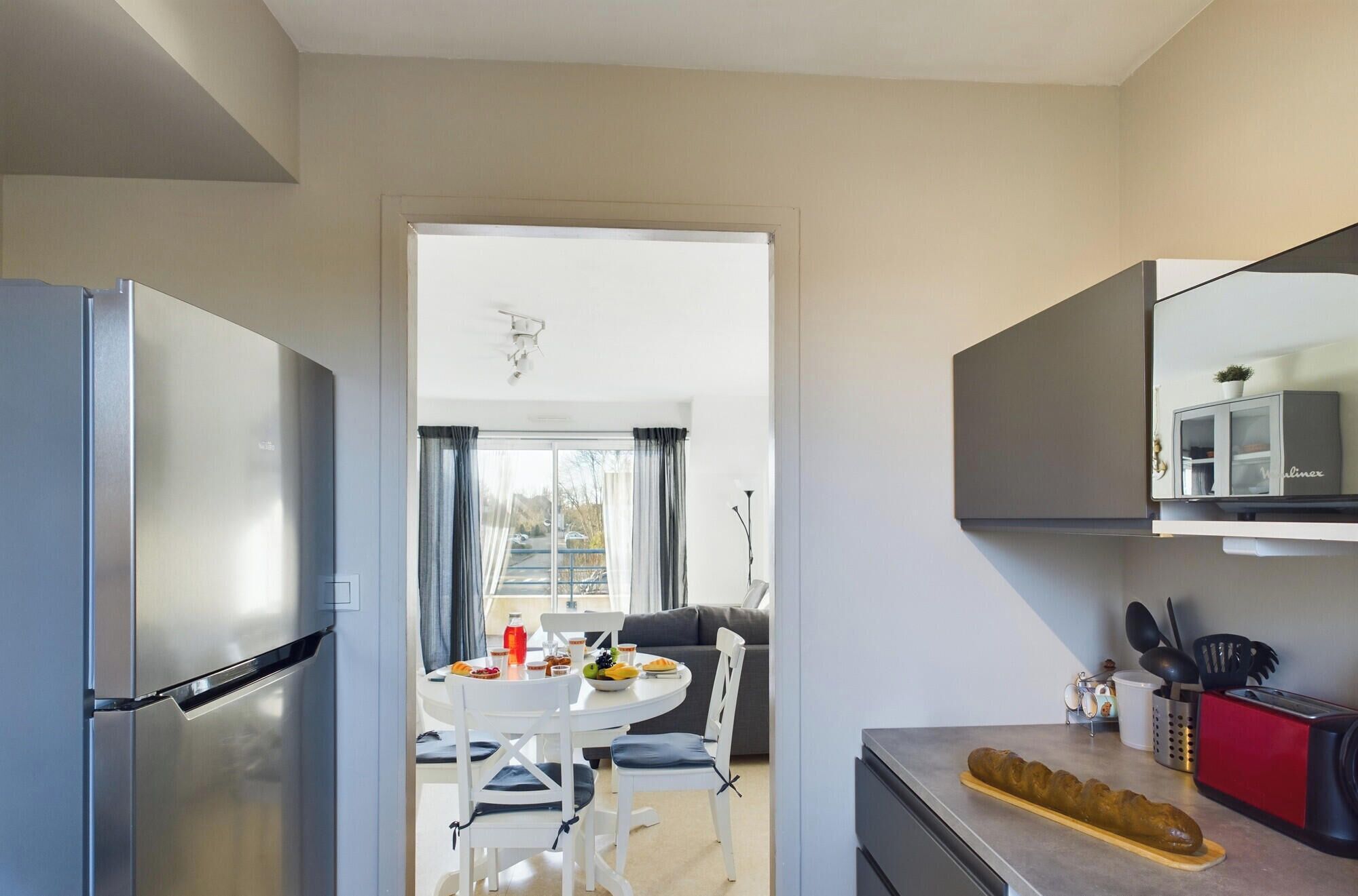 Une cuisine moderne équipée d’un réfrigérateur, d’une table à manger avec des chaises, d’un îlot central et d’une grande fenêtre.