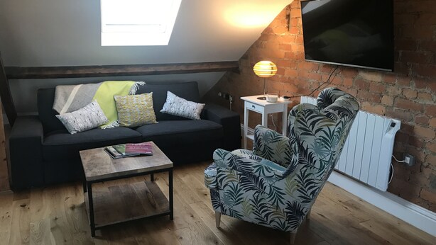 A cozy loft-style living room with a black sofa, patterned armchair, and a small wooden coffee table. A white side table with a lamp is against the brick wall, and a flat-screen TV is mounted above. - Windows Over Stroud Penthouse Apartment With Views (Stroud)