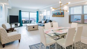 A modern living room with a glass dining table, white upholstered chairs, a sofa, and a television. - Regency Towers #708 West | Gulf Views, Pool! (Pensacola Beach)