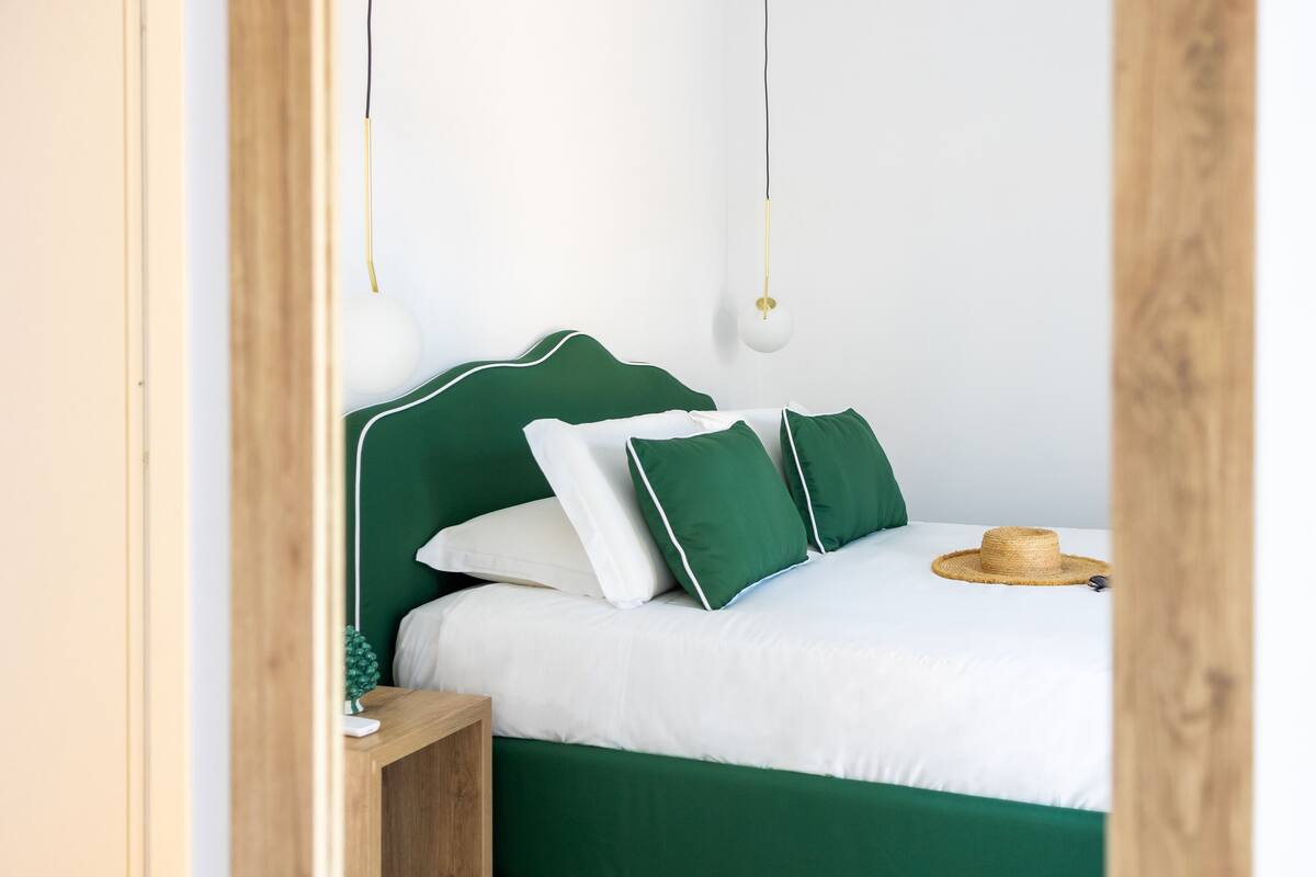 A neatly made bed with green bedding and white pillows, a small bedside table with a lamp, and a straw hat on the bed.