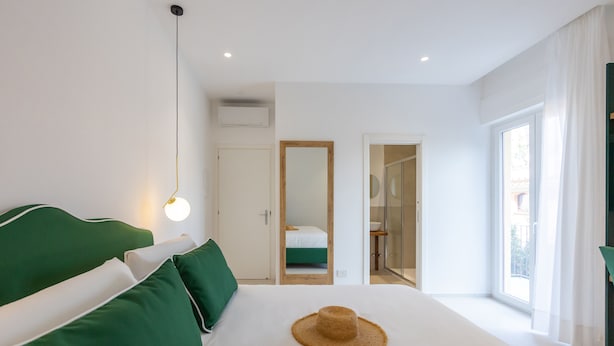 A modern hotel room with a large bed, green bedding, a straw hat on the bed, a bedside lamp, and a view of the city through large windows. - Vittoria Rooms Sorrento (Sorrento)