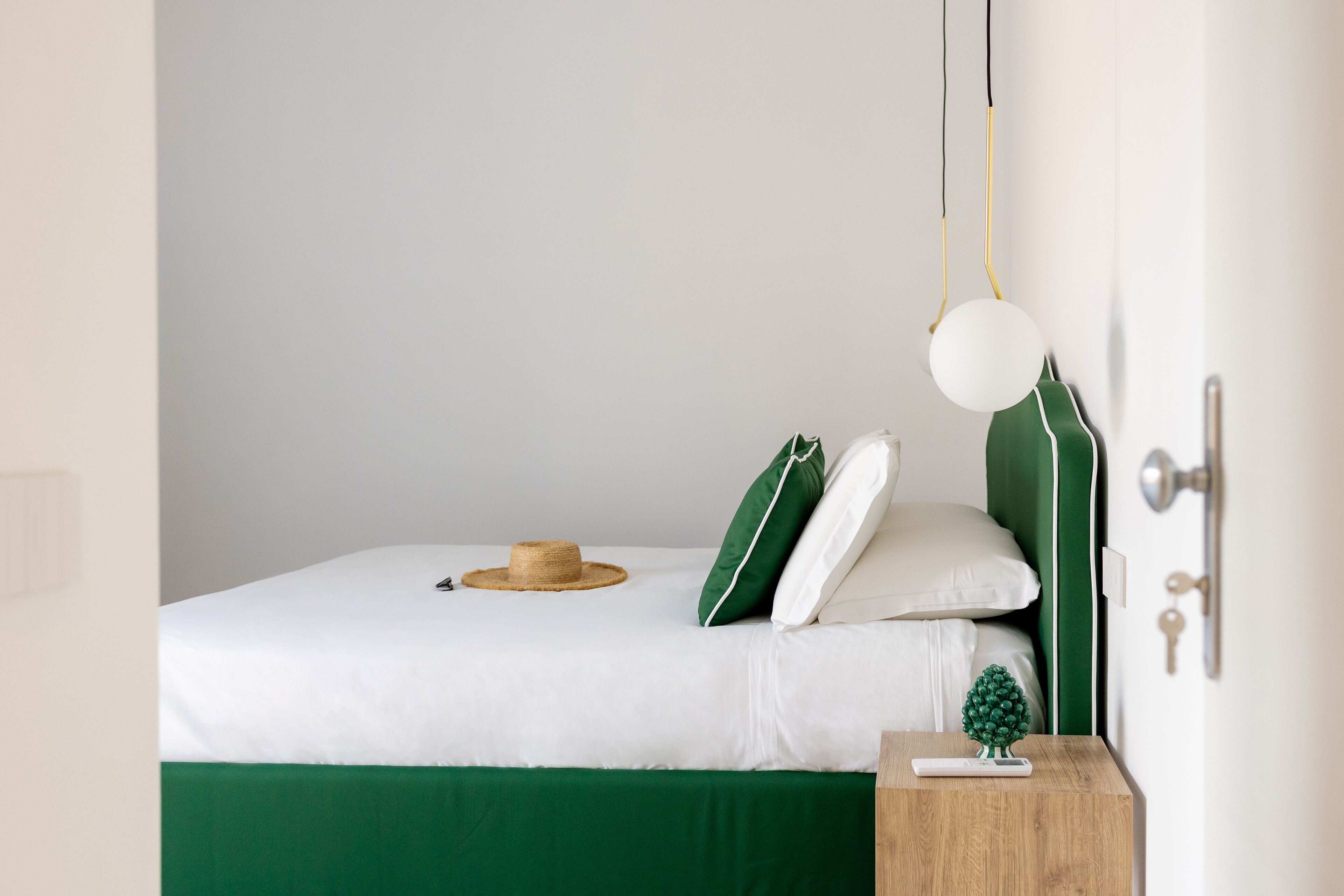 A neatly made bed with white bedding and green accents, a small wooden nightstand with a decorative item, and a simple hanging light fixture.