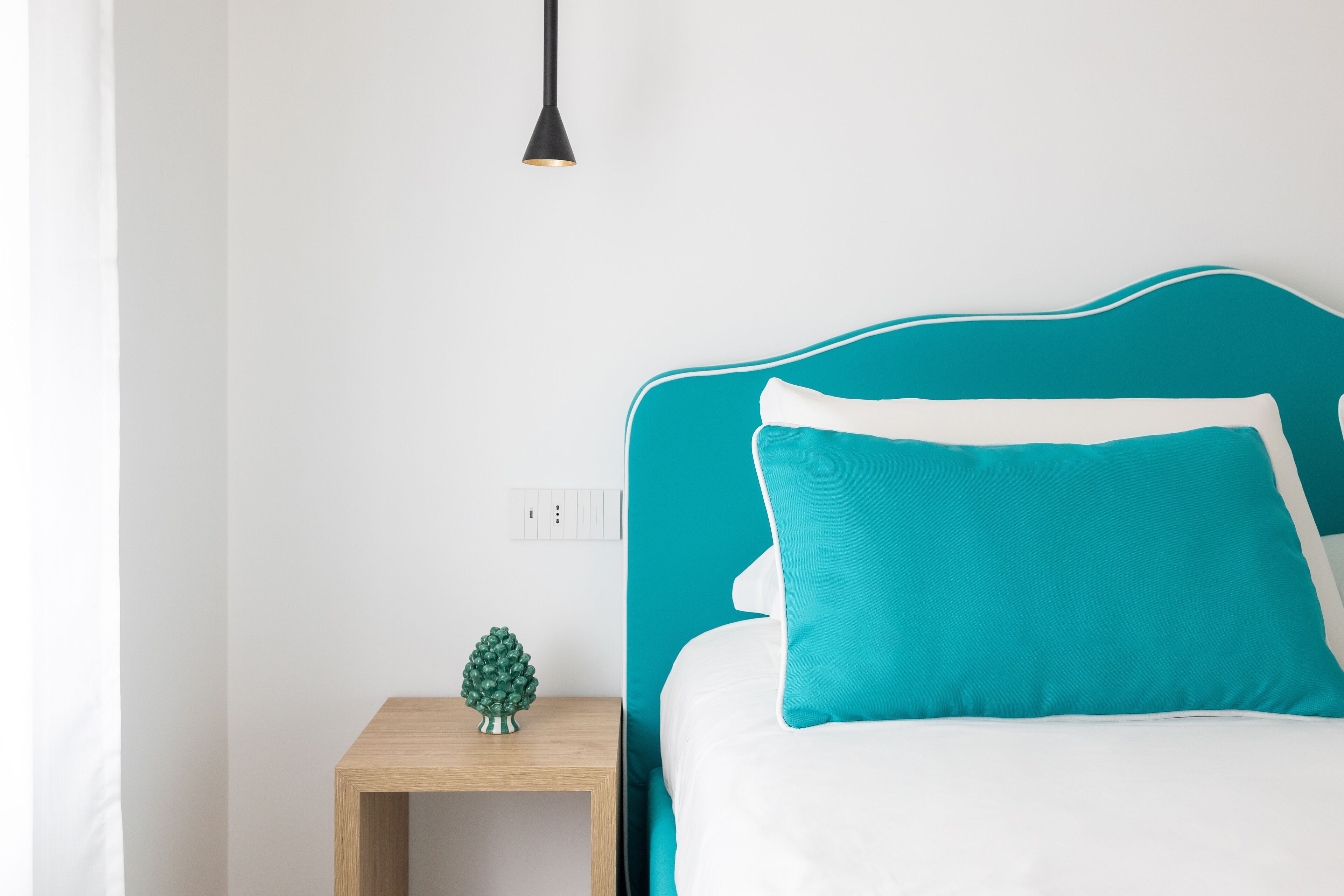 A neatly made bed with teal and white bedding, a small wooden nightstand with a green decorative item, and a black pendant light.