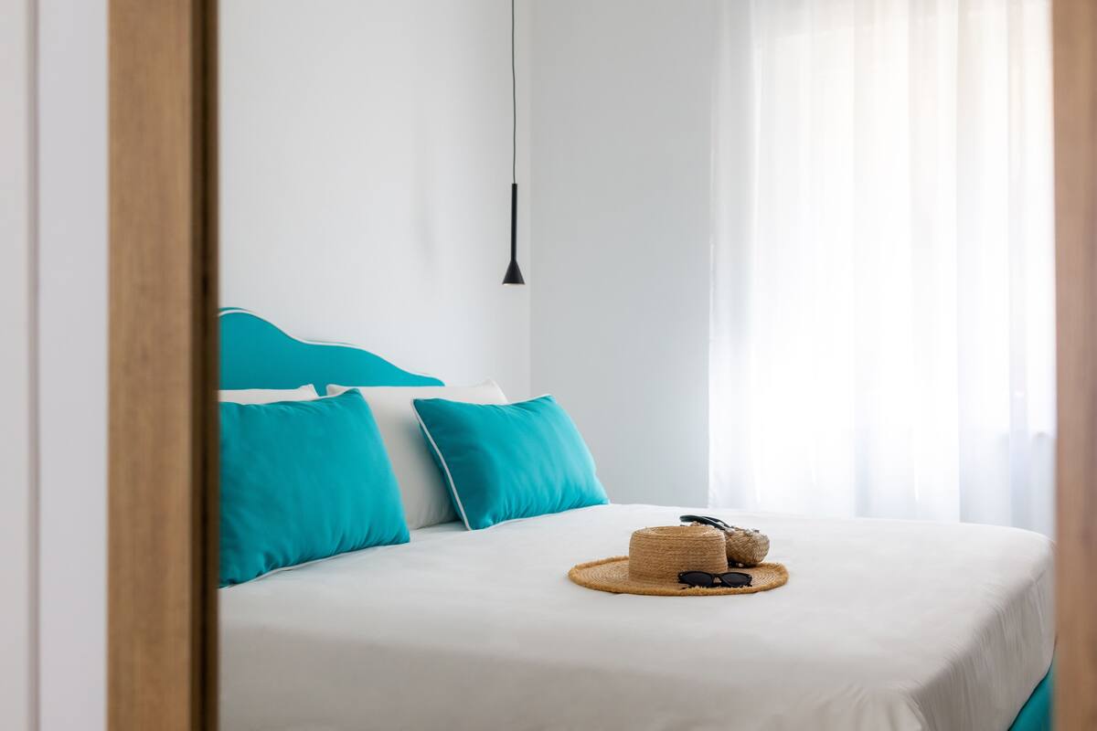 A neatly made bed with a straw hat and sunglasses on it, white bedding, and teal pillows.