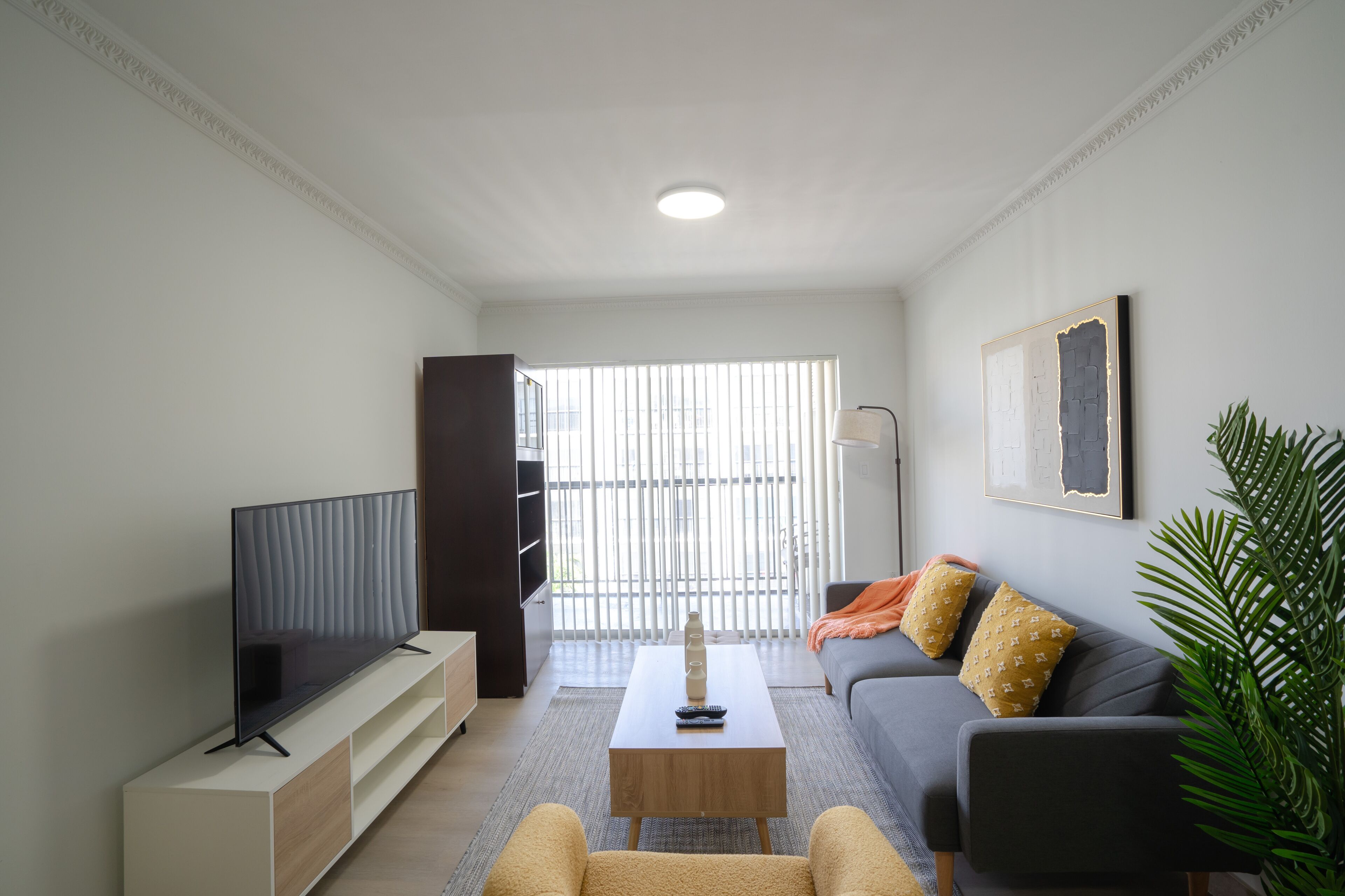 A modern living room with a flat-screen TV, a wooden coffee table, a grey sofa with patterned cushions, and a large window with blinds.