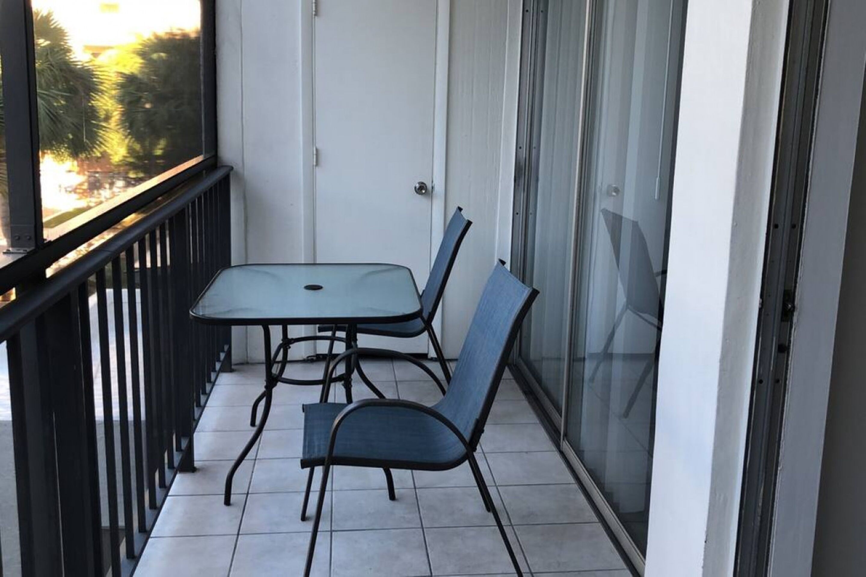 A balcony with a table and two chairs, a sliding glass door, and a view of palm trees.