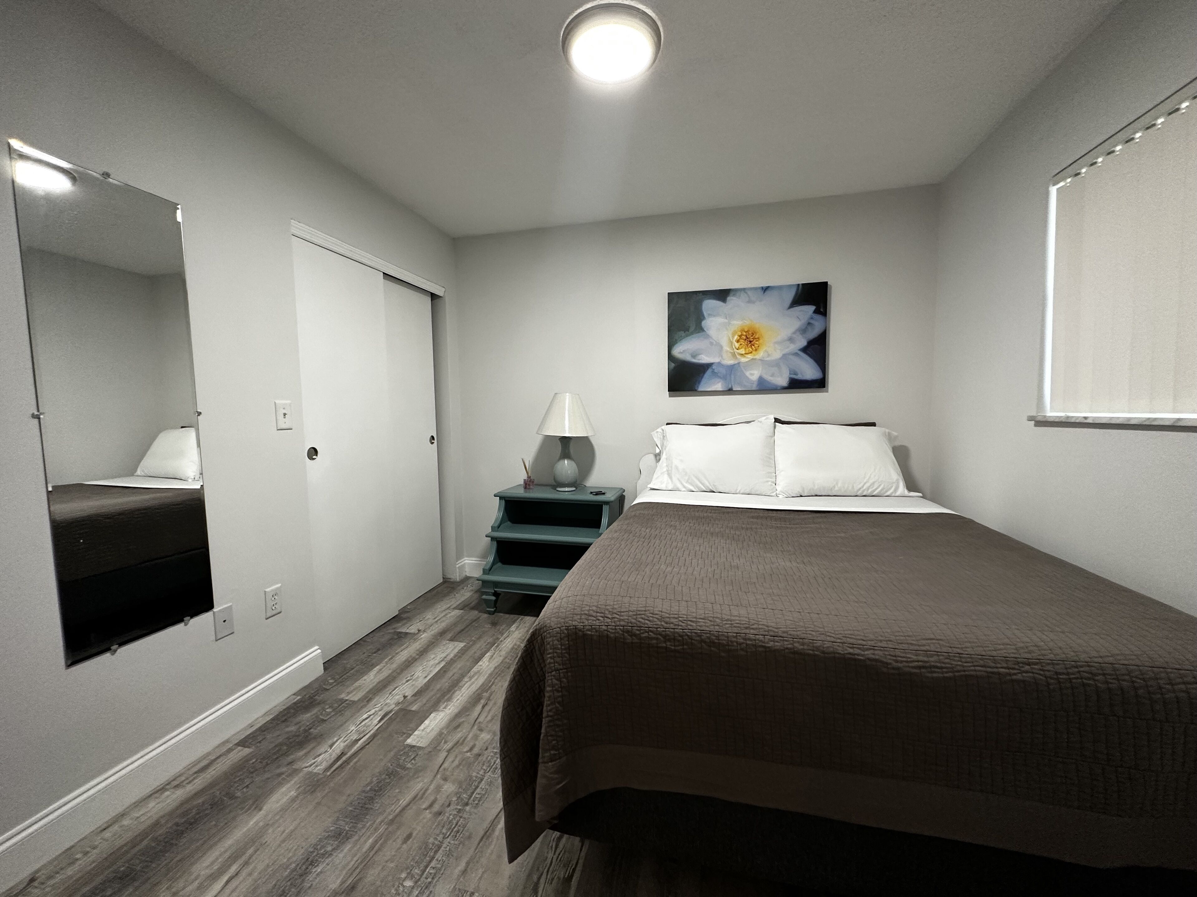 A bedroom with a bed, a nightstand with a lamp, a mirror, and a wall-mounted flower painting.