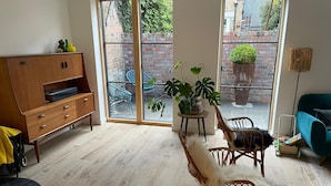 Un salon comprenant un canapé, des fauteuils, un meuble en bois et une grande fenêtre donnant sur un mur de briques et la verdure à l’extérieur.