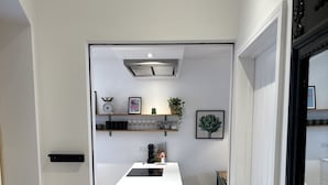 Une cuisine moderne avec des meubles blancs, un îlot central et un four encastré. Des  étagères au-dessus de l’îlot servent à ranger divers ustensiles de cuisine, et un tableau encadré y est également accroché.