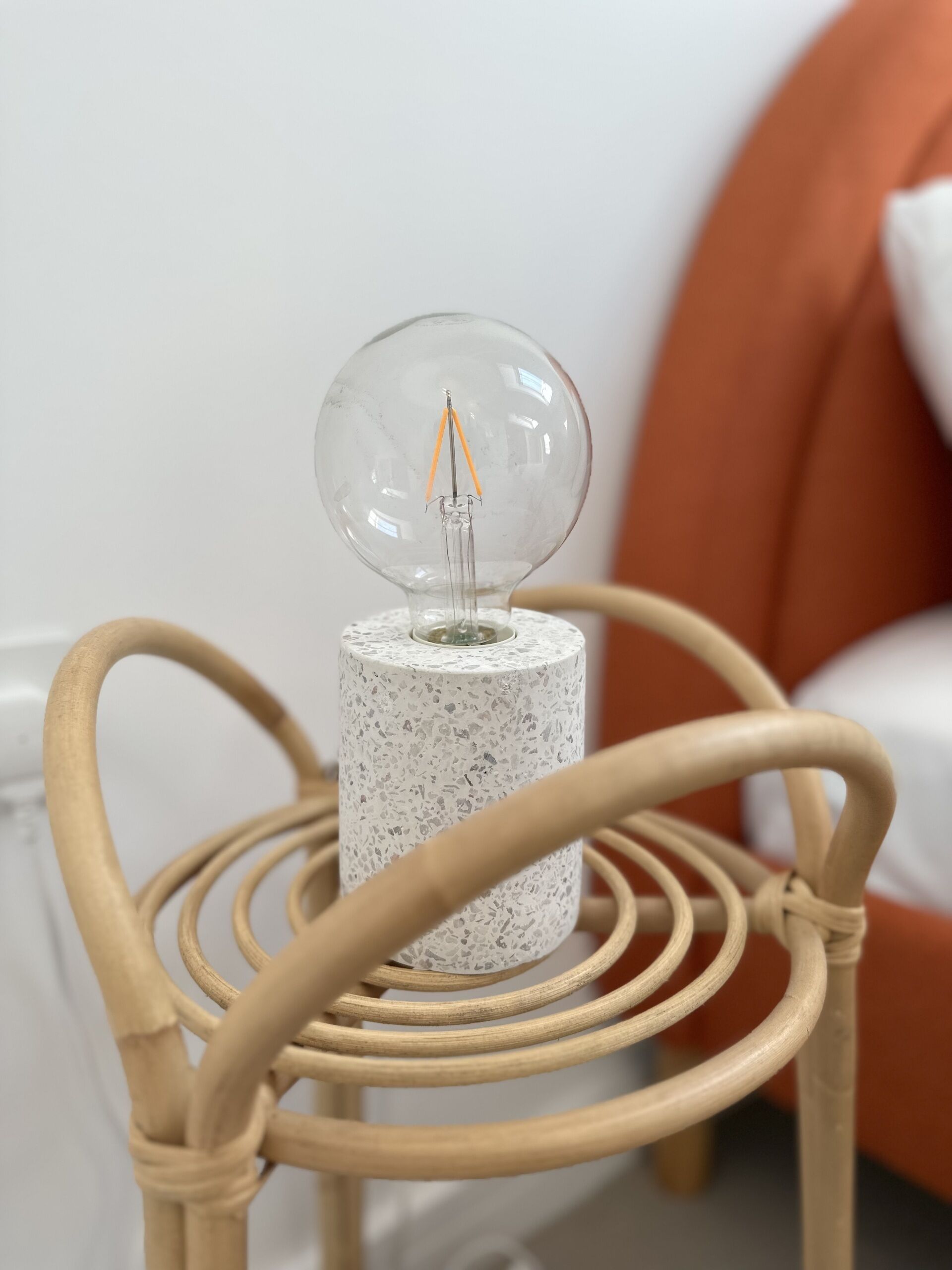 A clear glass light bulb on a speckled cylindrical base, placed on a woven chair with a white cushion in the background.