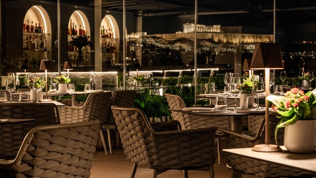 Un espacio exterior para comer con sillas y mesas de mimbre, una barra con estantes llenos de botellas y vistas a la ciudad de noche.