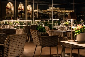 An outdoor dining area with wicker chairs and tables, a bar with shelves of bottles, and a view of the cityscape at night.