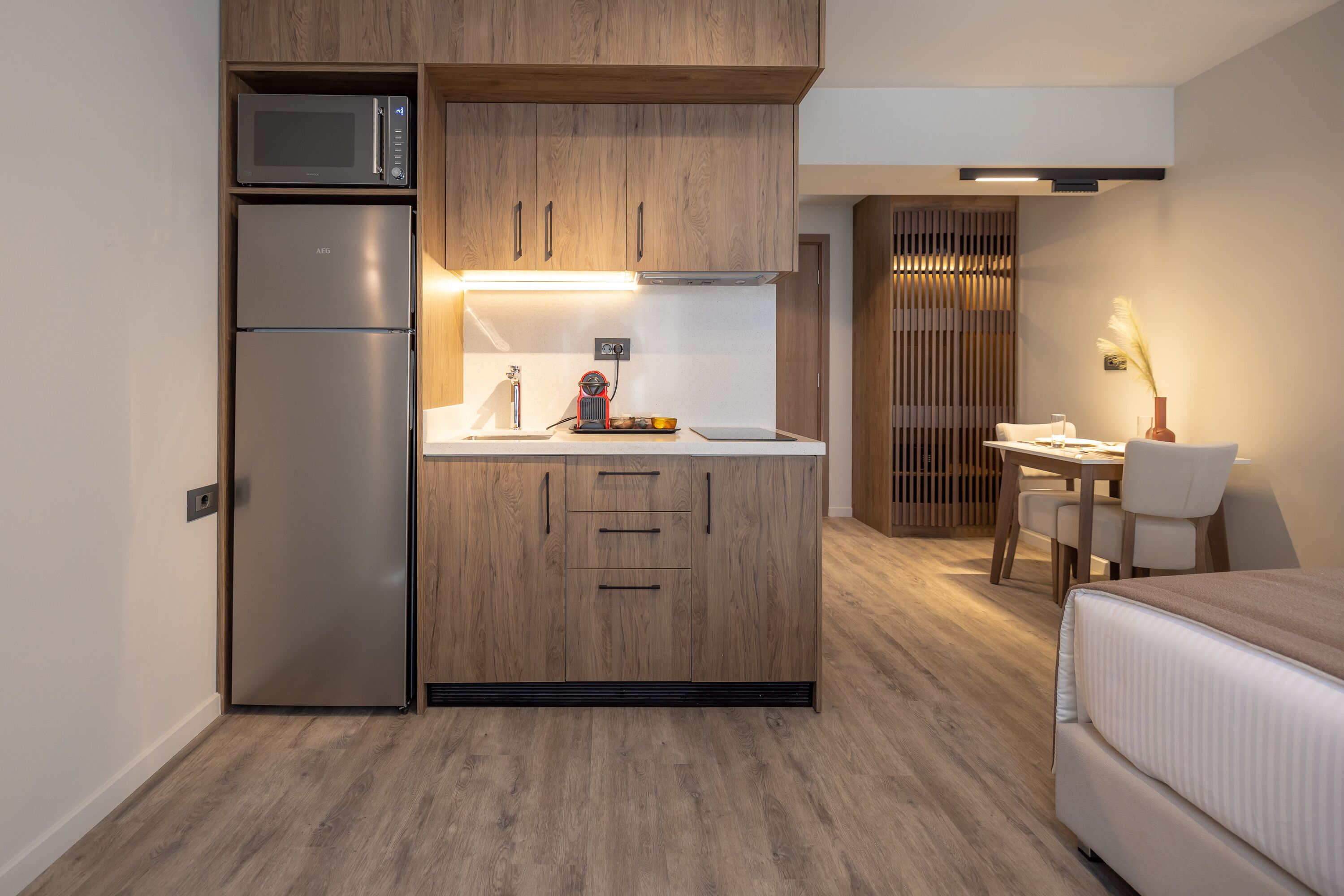 Una cocina compacta con microondas, refrigerador y armarios de madera, un pequeño comedor con una mesa y sillas, y una cama en la habitación contigua.