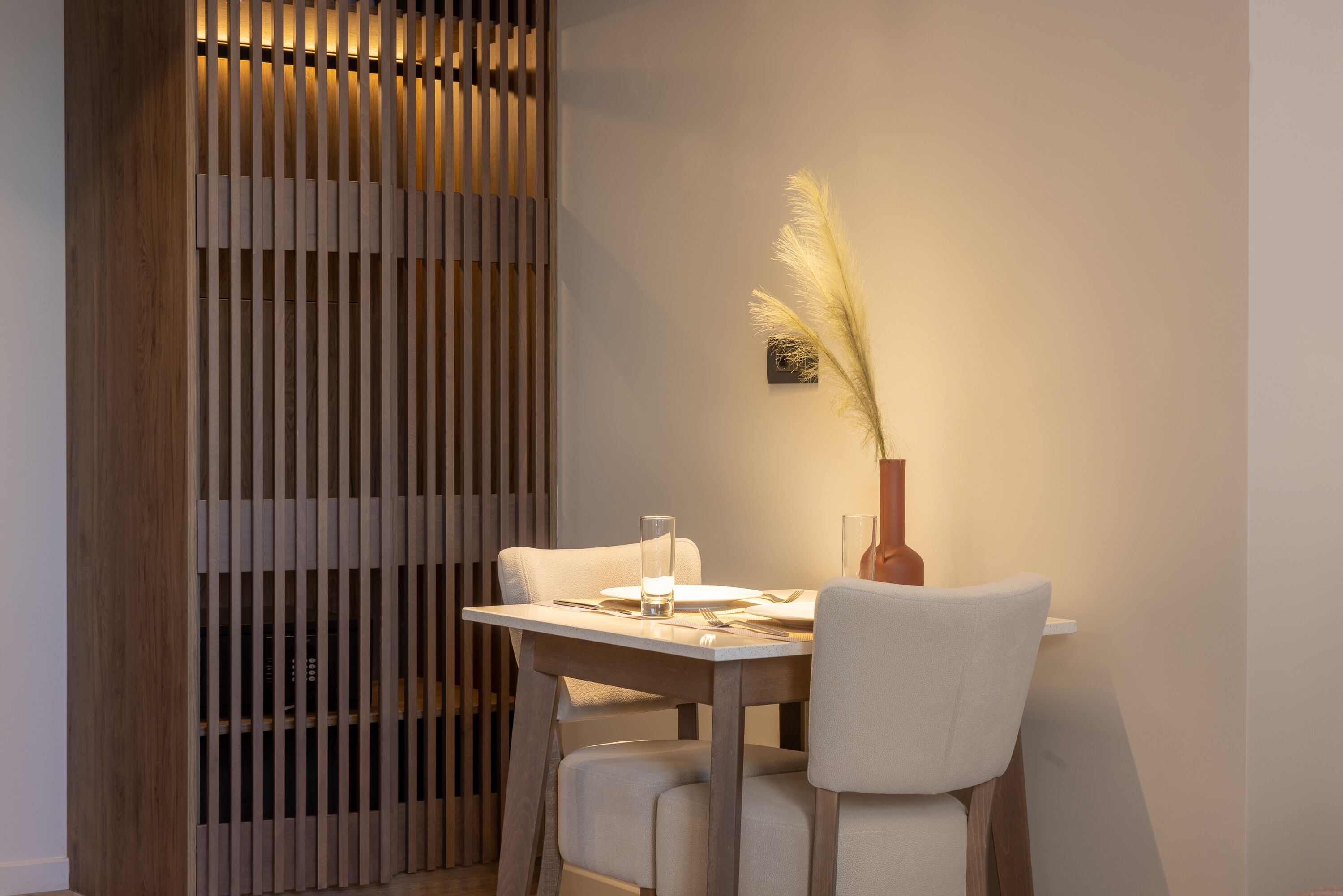 A dining area with a wooden partition, a table set with a glass, a vase with pampas grass, and two chairs.
