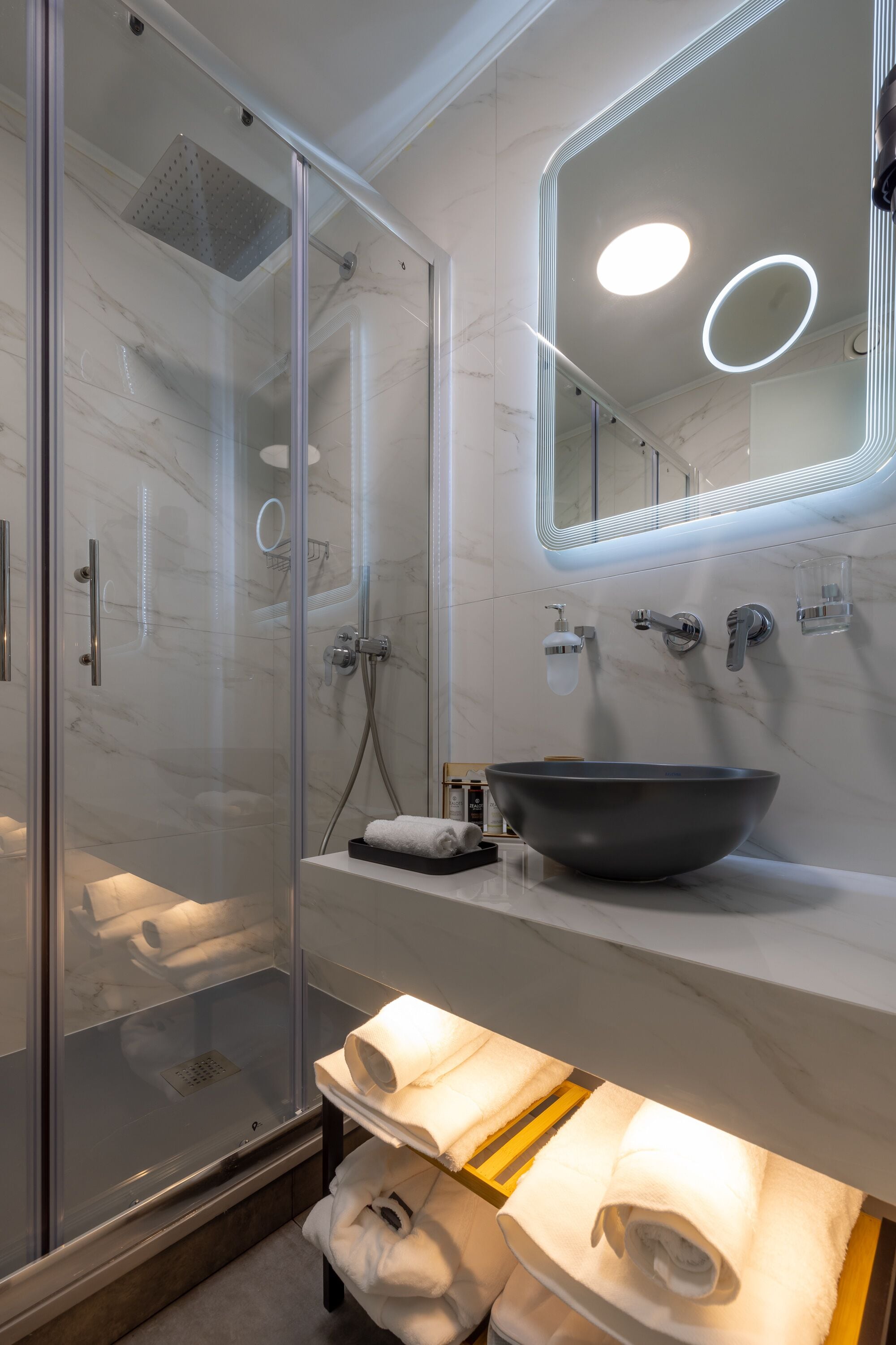 A modern bathroom with a glass-enclosed shower, a large mirror with LED lights, a marble sink, and neatly arranged towels.