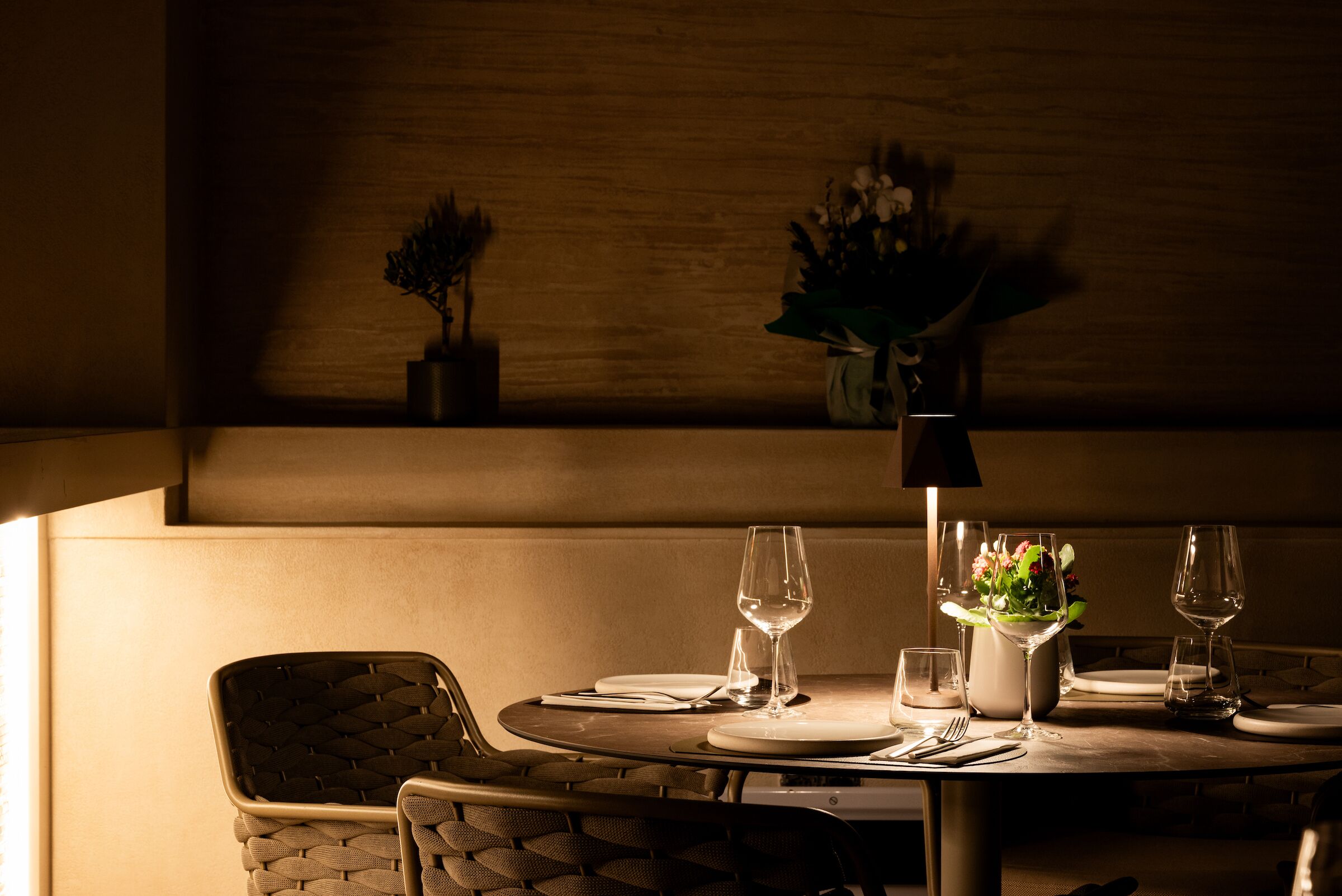 A dimly lit dining area with a round table set for two, accompanied by a potted plant and a vase of flowers.