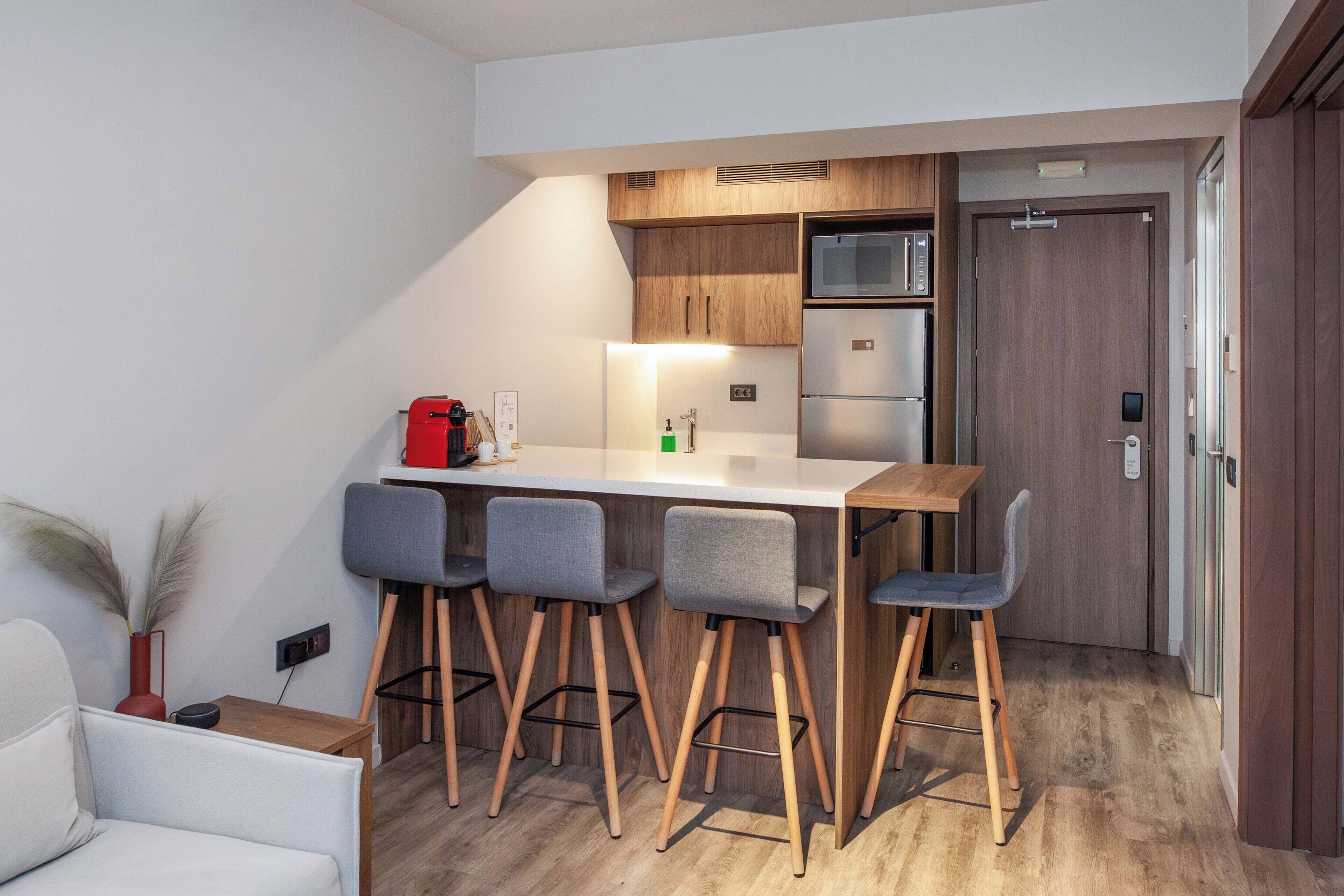 A compact kitchen with a white countertop, wooden cabinets, a stainless steel refrigerator, and a microwave. There are four grey bar stools and a white chair.