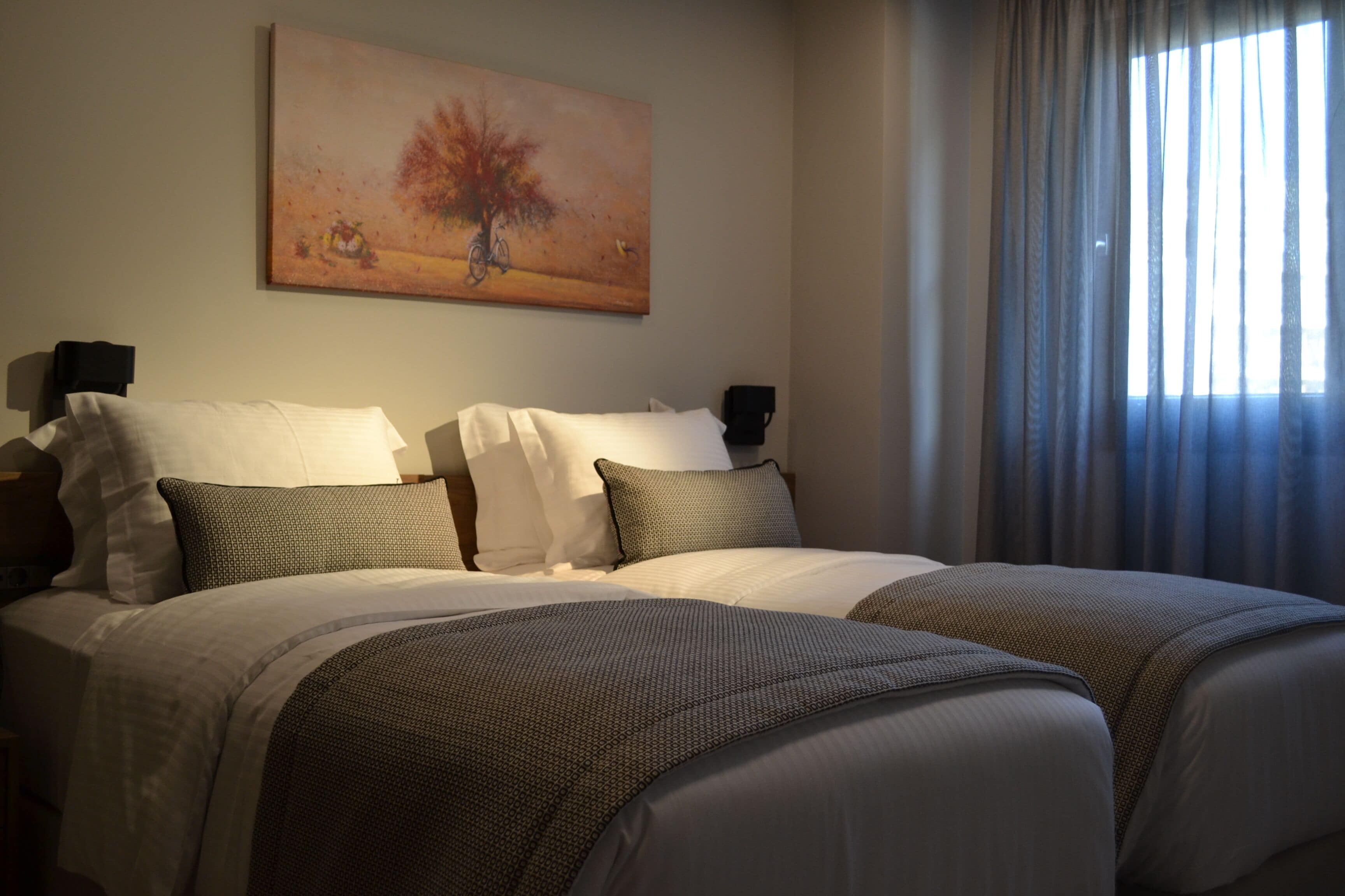 A hotel room with a large bed, two bedside tables, a painting on the wall, and a window with curtains.