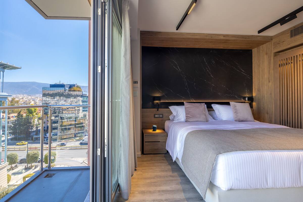 Une chambre avec un grand lit, une table de chevet, une tête de lit en bois et une vue sur le paysage urbain, visible à travers une grande fenêtre.