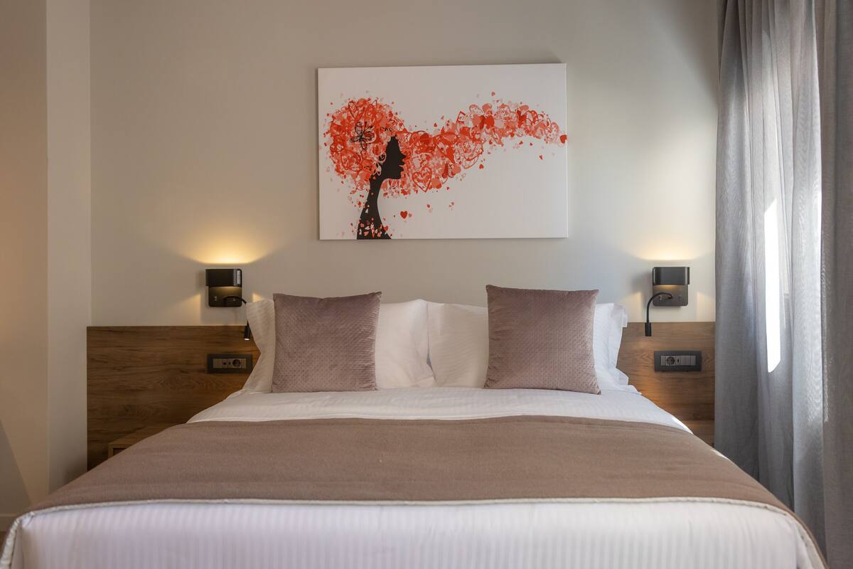 Une chambre d’hôtel avec un grand lit, deux tables de chevet avec des lampes, une œuvre d’art accrochée au mur et une fenêtre avec des rideaux.