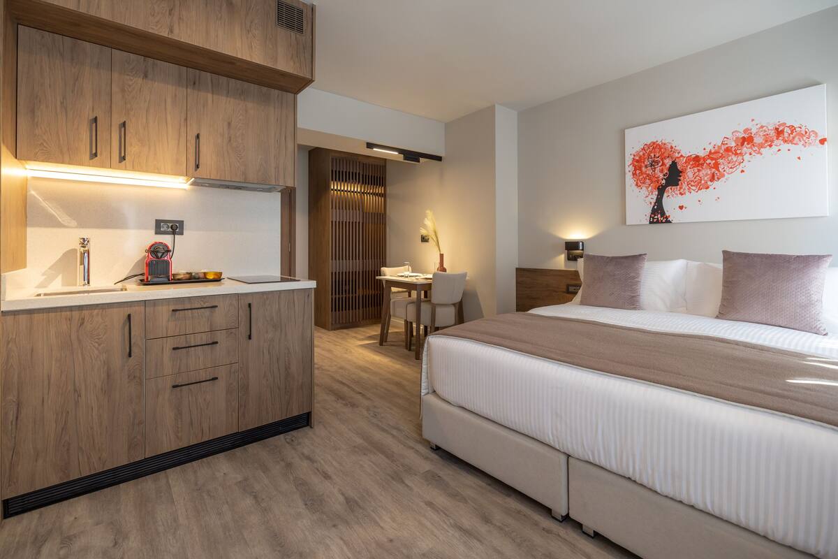 A modern hotel room with a kitchenette, a bed with a large window, and a painting on the wall.