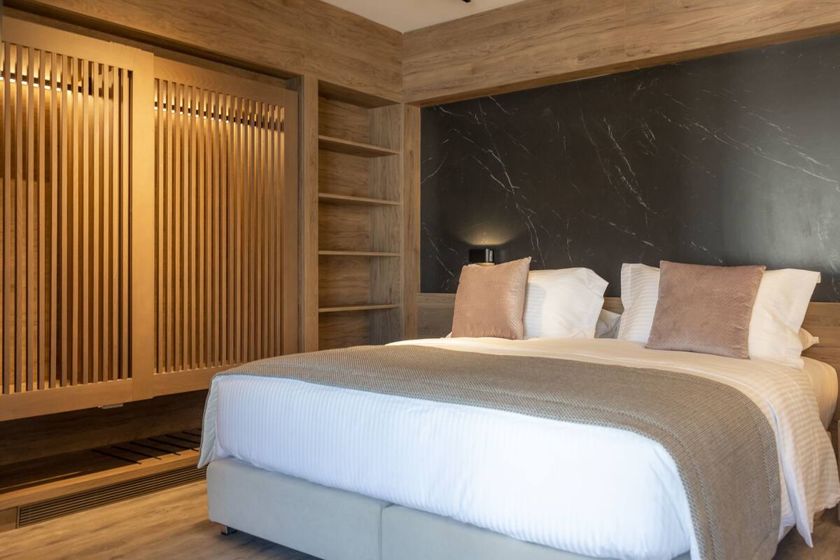 Une chambre à coucher moderne avec un grand lit, un lambris en bois, un mur d’accent en marbre et une table de chevet avec une lampe.