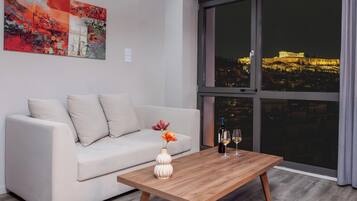 Una sala de estar moderna con un sofá, una mesa de centro de madera con un florero y copas de vino, y un amplio ventanal con vista a un paisaje urbano nocturno.