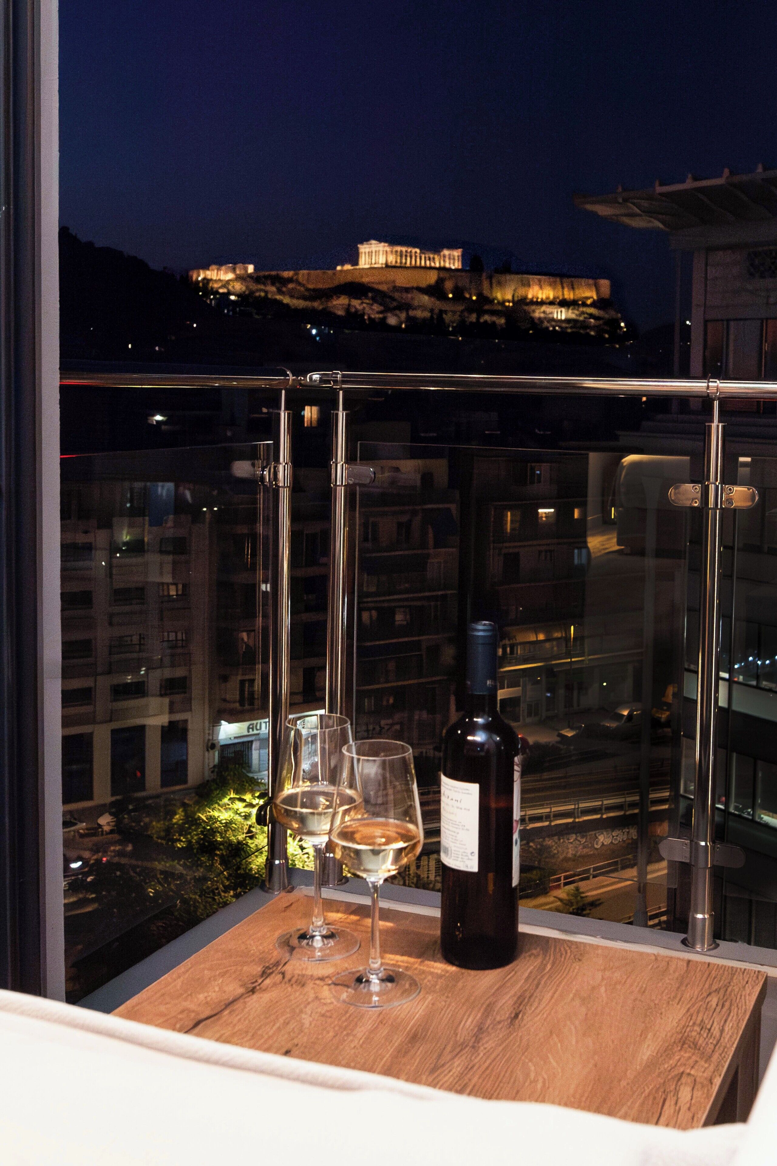 Un balcón con una botella de vino y dos copas, con vistas a un paisaje urbano donde destaca un edificio.