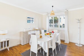 A dining area with a round table, white chairs, a fruit bowl, and a window with curtains.