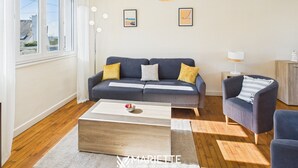 A modern living room with a grey sofa, a coffee table, and a blue armchair. There are two framed pictures on the wall, a floor lamp, and a window with curtains. - -HORTENSE- MAISON SABLES BLANCS Á 300M DES PLAGES (Concarneau)