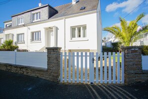 A two-story white house with a stone foundation, white picket fence, and a palm tree in the yard. - -HORTENSE- MAISON SABLES BLANCS Á 300M DES PLAGES (Concarneau)