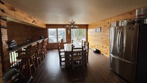 A wooden cabin kitchen with a dining table, chairs, a refrigerator, and a window overlooking a deck.