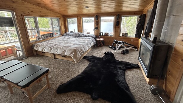 A cabin bedroom with a bed, a desk, a television, and a large window overlooking a lake.