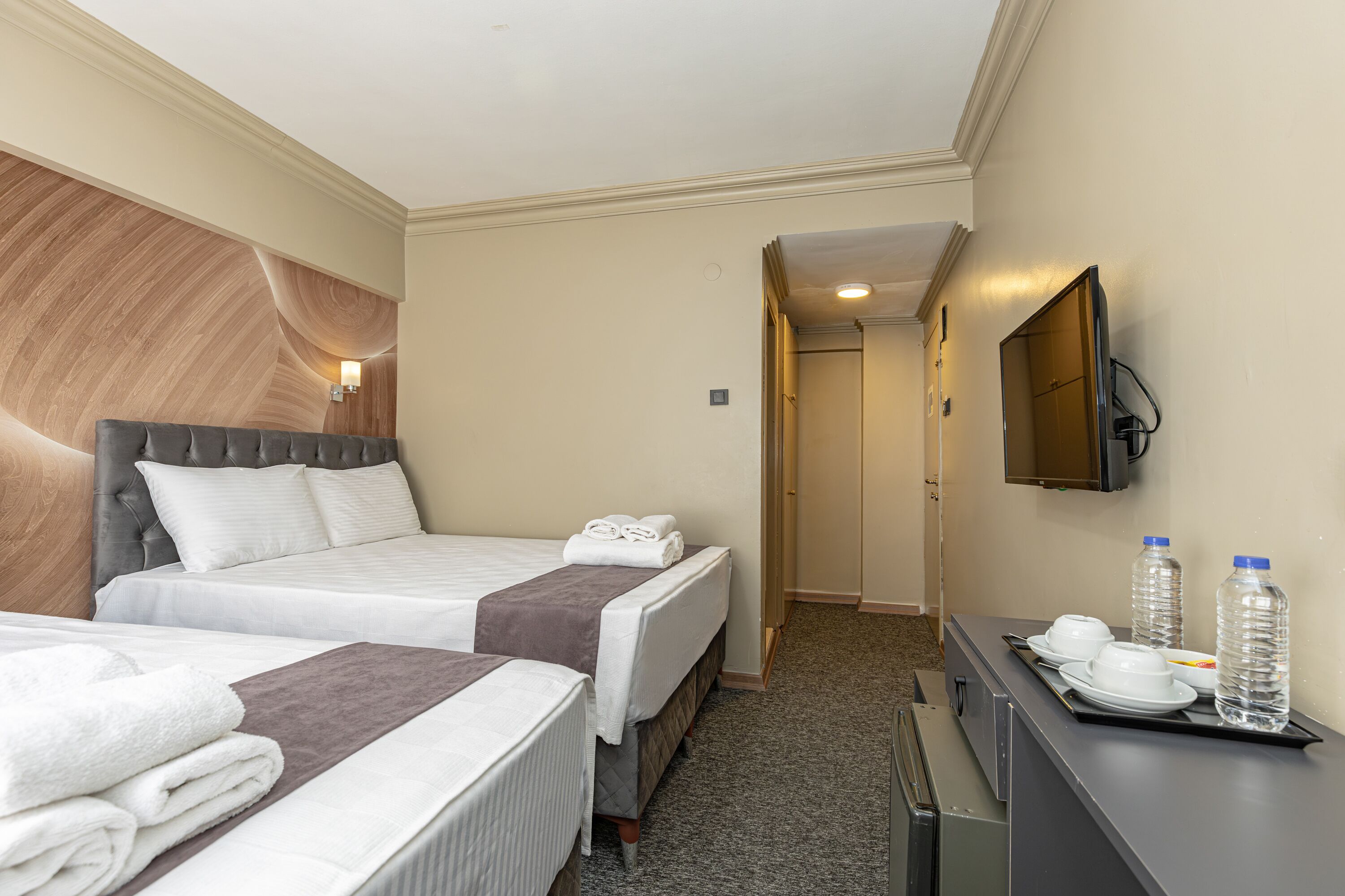 A hotel room with two beds, a flat-screen TV, a desk with a tray, and a water bottle.